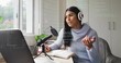 © vectorfusionart - Speaking woman in light blue sweater headset gesturing at laptop in home office with condenser mic