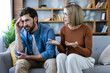 © Liubomir - Couple sitting on a couch in a living room having an argument, with the man looking upset and distressed, while the woman expresses her feelings with an open hand gesture