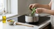© olga_demina - Hands adding fresh basil and thyme herbs to a boiling pot on an induction stove while preparing a healthy homemade meal with olive oil and vegetables in a kitchen