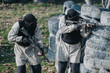 © qunica.com - Two masked players crouch behind a tire barricade, aiming their rifles in a paintball match.