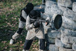 © qunica.com - Two players in camouflage protect their positions behind a tire barricade using paintball markers. An outdoor game scene with protective masks and gear.