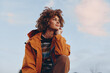 © SHOTPRIME STUDIO - A smiling woman with curly hair wearing a rainbow sweater and orange jacket looks thoughtfully into the distance outdoors. The woman embodies a positive lifestyle and casual fashion.