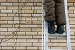 © Анатолий Еремин - Worker's legs in brown pants and black boots on a metal ladder against a beige brick wall. Black cables hang nearby, suggesting construction or repair. Industrial feel with ample copy space