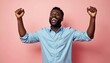 © Viktor - Happy Black man laughs with arms raised in victory, wearing a light blue shirt. He celebrates success or achievement against a soft pink studio background, expressing pure joy and excitement.