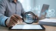 © Tabinda - A man examines a checklist on a magnifying glass while sitting at a desk with a tablet and laptop in a modern office setting.