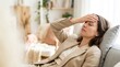 © ryker - Woman in beige suit resting on sofa with hand on forehead looking tired and pensive in bright living room