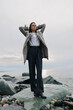 © SHOTPRIME STUDIO - Young woman stands on rocky shore wearing wide pants and coat, looking confident and relaxed with hands behind head. Cloudy sky and sea background create calm natural atmosphere.