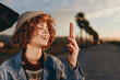 © SHOTPRIME STUDIO - Happy woman with curly hair smiling outdoors, wearing a rainbow sweater and denim jacket, holding smartphone, enjoying sunset, casual lifestyle, blurred road and palm trees background