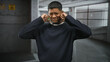 © Krakenimages.com - Man plugs ears with fingers and grimaces while standing in a dim concrete parking building, wearing a dark sweater and beard visible; annoyance.