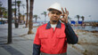 © Krakenimages.com - Hispanic man construction worker in red coveralls and white hard hat making ok sign with right hand on sunny beach promenade; pride competence.