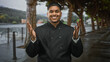 © Krakenimages.com - Hispanic man chef in black uniform smiles with open palms beside riverside railing on a street promenade; welcome hospitality joy.