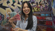 © Krakenimages.com - Woman holding yuan banknotes outdoors on graffiti-adorned street showcasing vibrant urban culture and economy with smiling expression and casual attire contrasted against artistic backdrop.