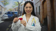 © Krakenimages.com - Young woman doctor with stethoscope holding red heart on urban street outdoors with confident expression wearing medical uniform promoting health awareness in community setting