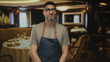 © Krakenimages.com - Man grimaces while wearing denim apron and glasses in a restaurant building dining room with set table behind him; resignation fatigue.