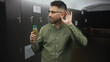 © Krakenimages.com - Hispanic man with beard holding green juice bottle while cupping his ear in a locker room; curiosity wellness routine.
