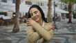 © Krakenimages.com - Confident hispanic woman smiling warmly on a lively street, wearing a beige blazer, embracing herself outdoors amidst palm trees and blurred cafes, showcasing happiness and serenity.