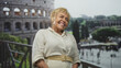 © Krakenimages.com - Senior hispanic woman smiling, face turned and head tilted, hands on railing at historic building colosseum in rome; travel joy.
