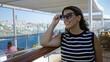 © Krakenimages.com - Young brunette woman lifts sunglasses and smiles, arm resting on wooden deck railing, hair blown by breeze, on ship building terrace with ocean view; serenity.