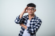 © Daniel - Young Asian man wearing round glasses, plaid shirt, and white tank top holds his head with one hand while grimacing in pain against a light gray background
