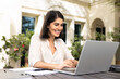 © fizkes - Happy young Latin freelancer woman working on online business project on home terrace, typing on laptop. Digital nomad using computer for remote work from outside
