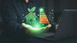 © shahabudin - A glowing green hand presents a digital icon of a sustainable house with leaves and an energy efficiency rating scale A to G alongside a laptop in a d