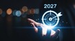 © simran - a hand is holding a glowing target with an arrow in it and the year 2027 above it. the background is dark with blurred lights.