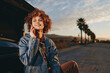 © SHOTPRIME STUDIO - A smiling woman in a rainbow sweater and denim jacket talks on a smartphone while sitting on the open trunk of a car during sunset on a palm tree lined road. Lifestyle concept.