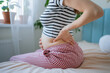 © wifesun - Pregnant woman feeling back pain, sitting with one hand supporting her baby bump and the other hand pressing against her lower back, illustrating common discomfort and health issues during maternity