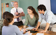 © JackF - Students solve problems together at table in classroom, teacher, standing in background, monitors process