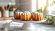 © Birul - Delicious Mini Bundt Cakes with Glaze on a White Pedestal in a Bright Kitchen.