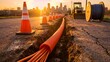 © CHLOÉ ANNE-MARIE - Sunlight illuminates a trench excavation site where underground fiber optic cable conduit is being installed near a construction vehicle and traffic cones against a city skyline.
