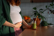 © wifesun - Pregnant woman holding her belly, standing in a kitchen and holding a vibrant green smoothie with a straw, focusing on healthy nutrition, vitamins, and a detox diet