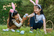 © geargodz - two happy child girl wearing bunny ears playing together and hunting for Easter eggs into basket on green grass in garden