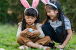 © geargodz - two happy child girl wearing bunny ears playing together and hunting for Easter eggs into basket on green grass in garden
