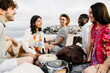 © Xavier Lorenzo - Multiracial group of young friends relaxes on a beach, engaging in conversation and playing drums, capturing the essence of a joyful summer vacation