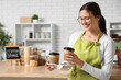 © Pixel-Shot - Happy young female barista with paper cups of coffee in cafe