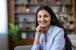 © Liubomir - Young adult indian woman smiling. Looking at camera with confidence and professionalism. Sitting in a domestic interior with a bookshelf background. Representing work from home and smart casual style