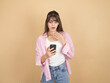 © Designerant - Shocked young woman looking at smartphone while covering her mouth with hand, wearing pink shirt and denim, standing against beige studio background with wide copy space. Surprise message concept.