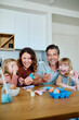 © we.bond.creations - Happy family looking at camera while decorating Easter eggs at a modern kitchen table, celebrating springtime holiday tradition