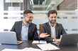 © Liubomir - Two smiling professional businessmen are discussing work and collaborating on a project, reviewing data on an open laptop during a meeting in a contemporary corporate office