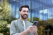 © Liubomir - Bearded man smiling and interacting with his smartphone, standing outdoors against a backdrop of a blurred corporate glass building, representing urban lifestyle and digital communication