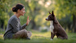 © dinatychynska - Woman trainer hand signal dog sitting obedience park grass, brunette sweater canine command session, pet bond training lifestyle green bokeh outdoor portrait.
