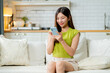 © Yingyaipumi - Happy young asian woman using mobile apps in living room,Smiling young asian female using smartphone sitting on couch at home.Messaging,Online Shopping,celebrate success,chatting online,Social Media