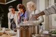 © AnnaStills - Caucasian chef stirring pot while three middle aged Caucasian women observing and smelling ingredients during cooking class, steam rising from pot, kitchen counter with food visible