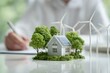 © Marina Demidiuk - Energy-efficient house model with green tree and wind turbines. On a background is an engineer working at desk. Eco-friendly living and modern technology. Sustainable home, advanced building materials