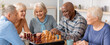 © Prostock-studio - A group of five seniors gathers around a chessboard in a bright indoor space. They are engaged in a game of chess, sharing smiles and drinks. The atmosphere is lively.