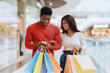 © Prostock-studio - Two young people walk through a mall smiling and holding shopping bags. One person checks a phone while the other looks at them. They enjoy their shopping time together.