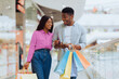 © Prostock-studio - A man and woman walk inside a shopping mall holding bags. They smile at each other as they check their phone. The atmosphere is lively with shoppers around them.
