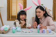 © geargodz - mother and preschool child girl wearing bunny ears painting and decorating egg  together at home on easter day. happy family holiday activity concept