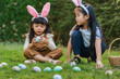 © geargodz - two child girl wearing bunny ears playing together and hunting for Easter eggs into basket on green grass in garden